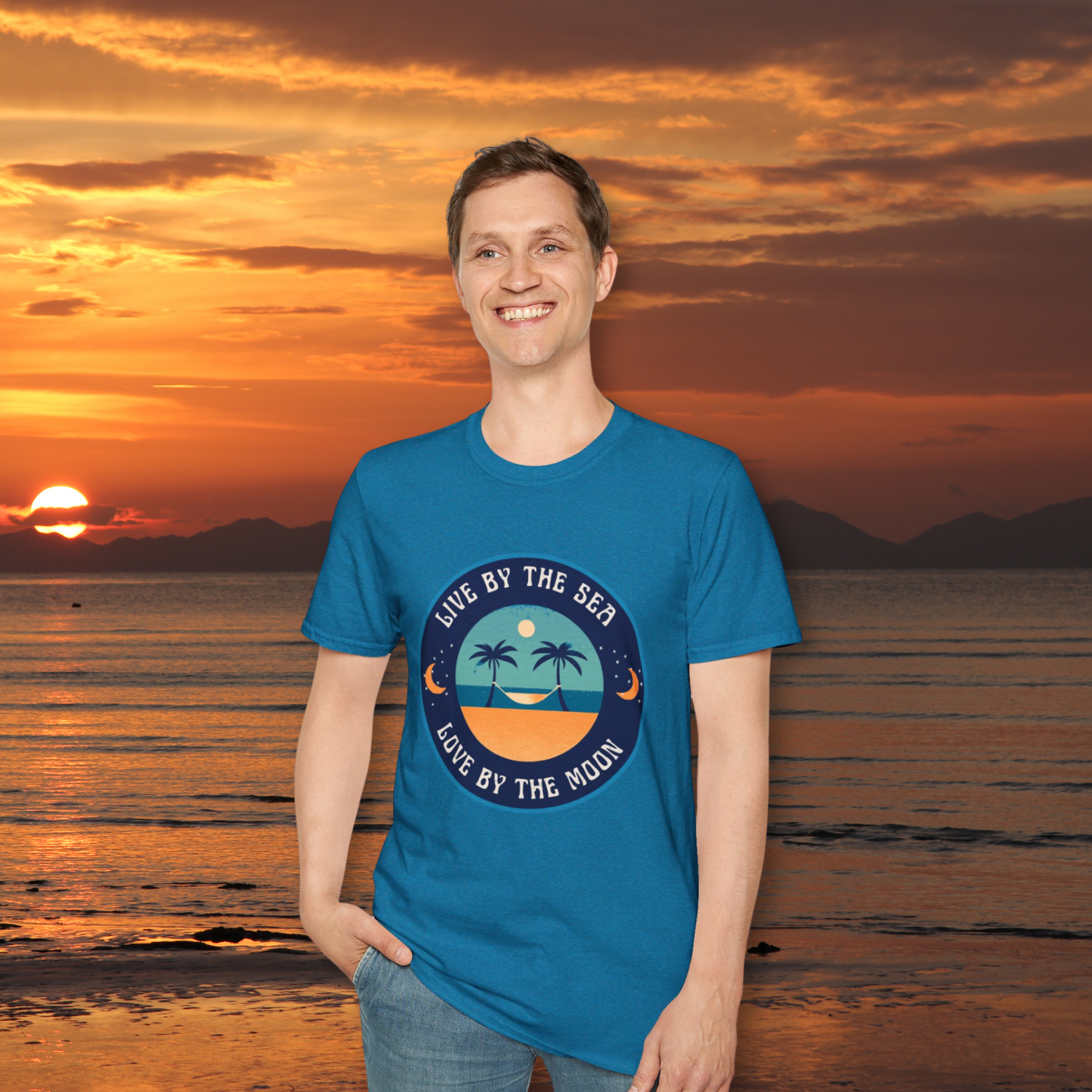 Man wearing Live By The Sea Love By The Moon Tee Shirt with a sun setting on the ocean in the background.